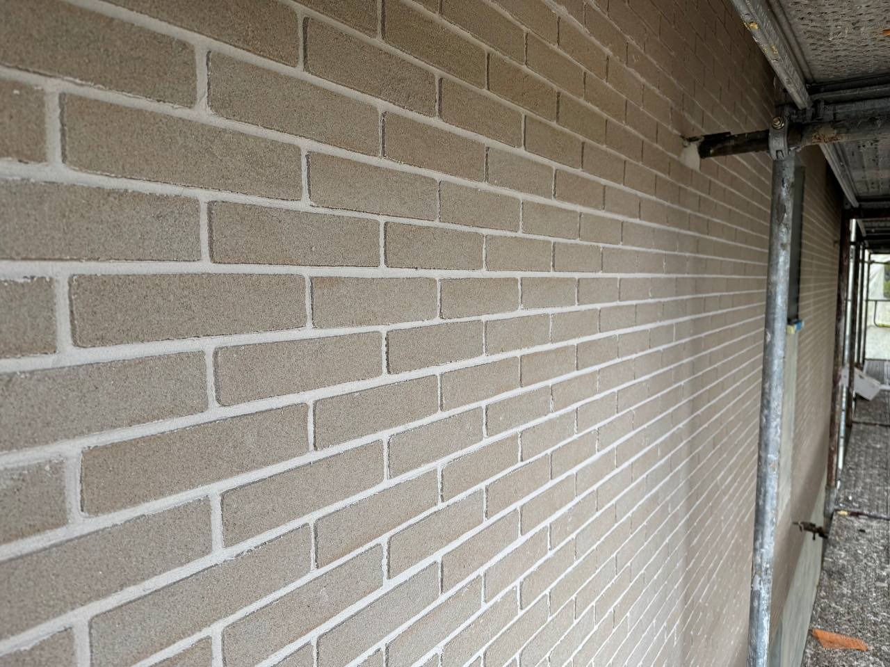 Ściana z cegieł klinkierowych w odcieniach beżu, z widocznymi białymi fugami, sfotografowana pod kątem z rusztowaniem w tle.