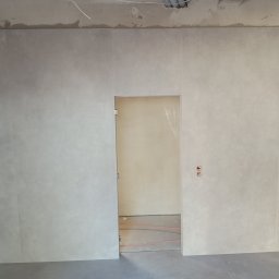 Dmytro Kornilov - Wykończona ściana w łazience z dużych, beżowych płytek, widoczne gniazdka i fragment instalacji podtynkowej. W tle widoczne ogrzewanie podłogowe.
