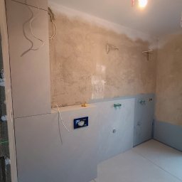 Dmytro Kornilov - Wnętrze łazienki w trakcie prac wykończeniowych: ułożone płytki podłogowe i częściowo ścienne, widoczne instalacje wodne i elektryczne, surowa ściana w tle.