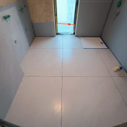 Dmytro Kornilov - Jasne, kwadratowe płytki podłogowe w trakcie układania w łazience z oknem dachowym. Widoczne przygotowane ściany i instalacje.