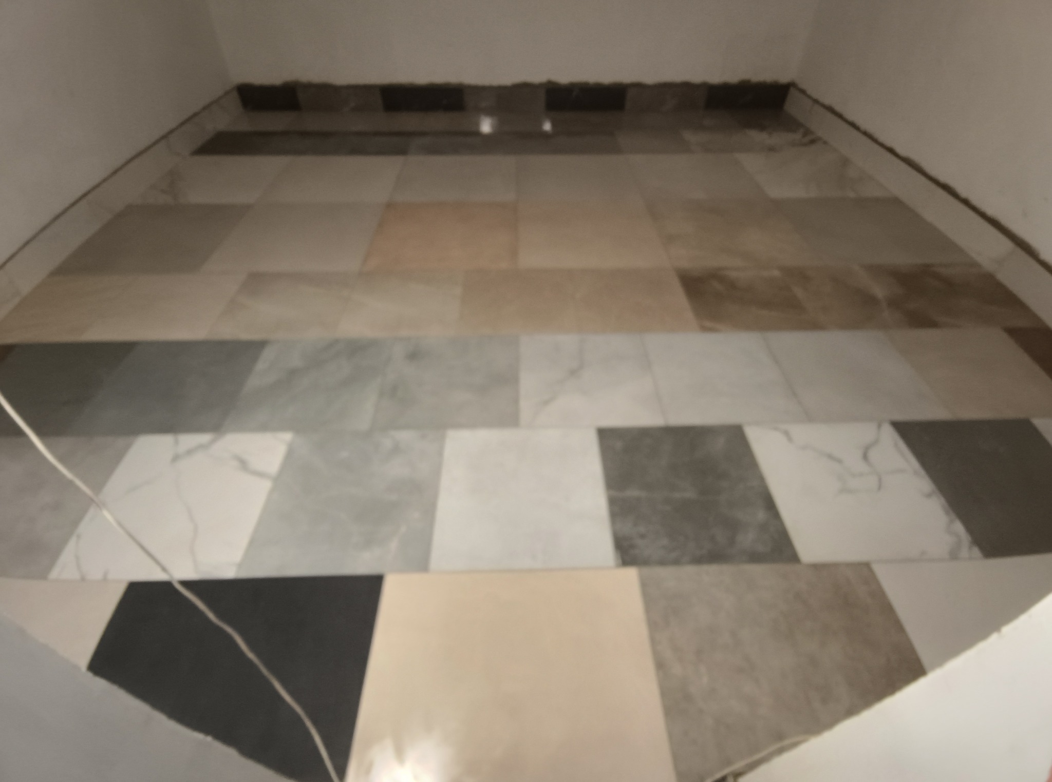 Ułożona podłoga z płytek w różnych odcieniach szarości, beżu i bieli. Widoczne niedokończone wykończenia przy ścianach. Płytki ułożone w regularny wzór.