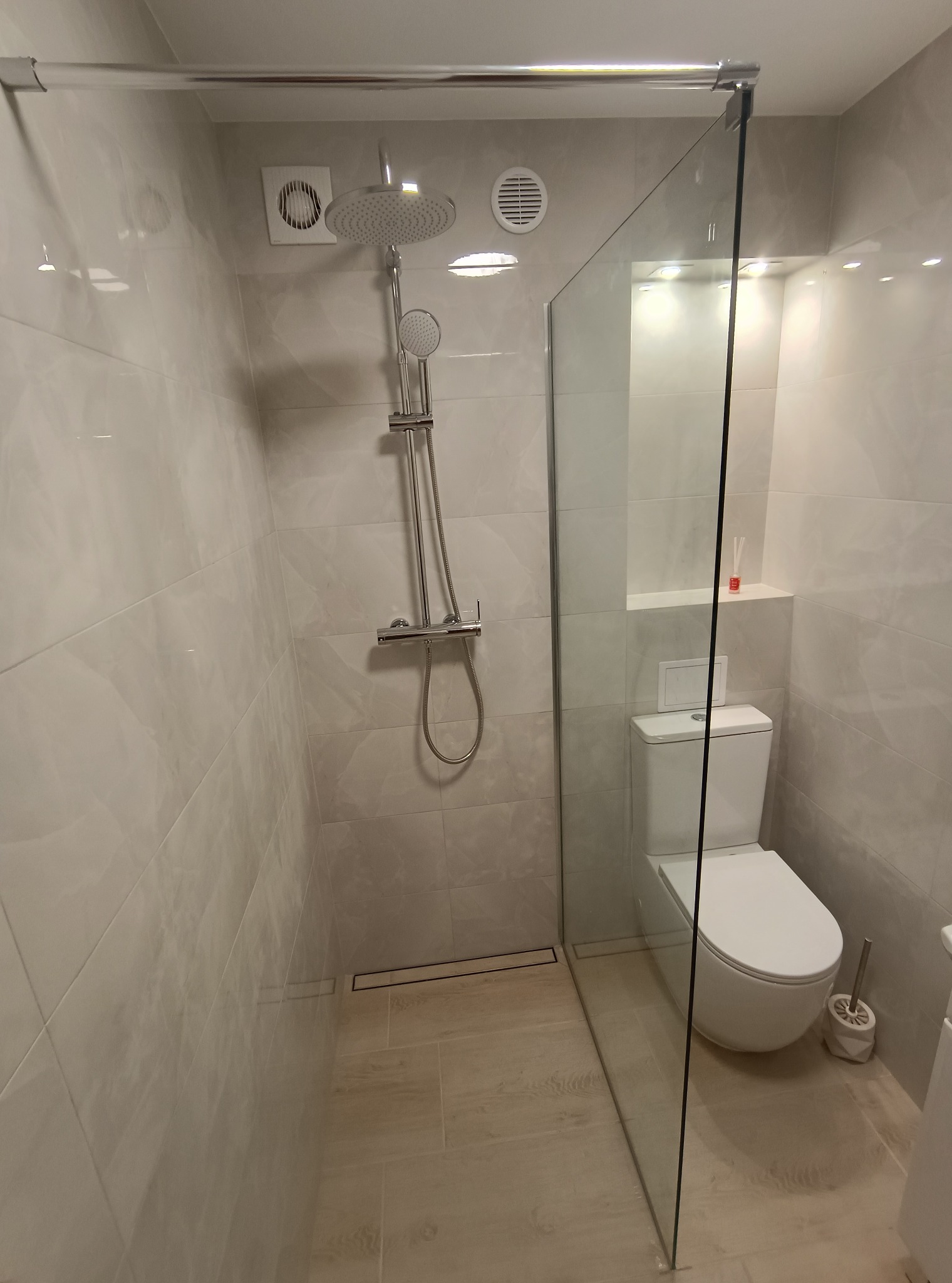 Jasna łazienka z kabiną typu walk-in, beżowe płytki imitujące drewno, toaleta w zabudowie i chromowana armatura. Minimalistyczny design z wentylacją.