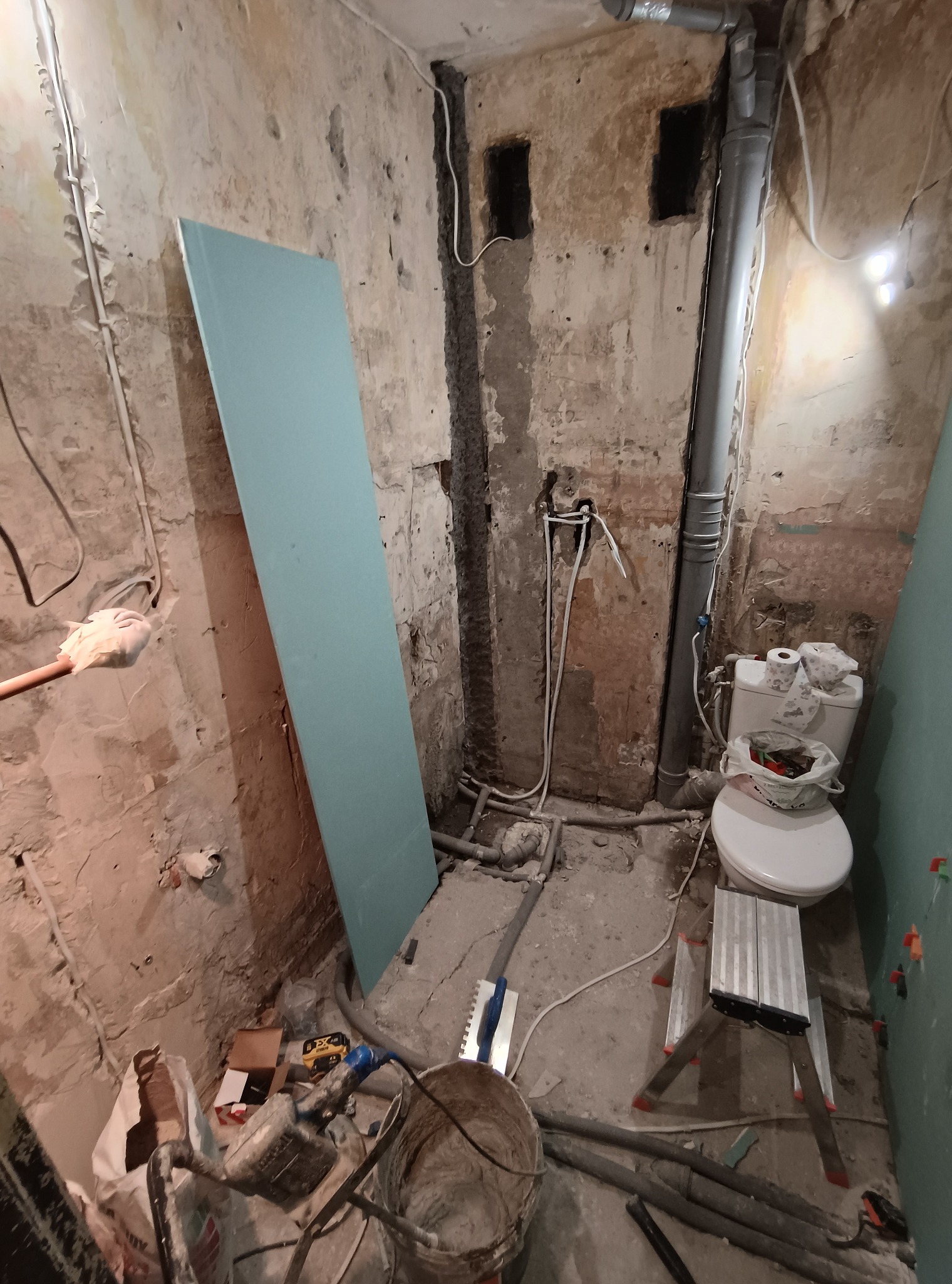 Wnętrze łazienki w trakcie remontu: surowe ściany, instalacje, płyta gipsowa oparta o ścianę, drabina i narzędzia. Widoczne rury i elementy instalacji sanitarnej.