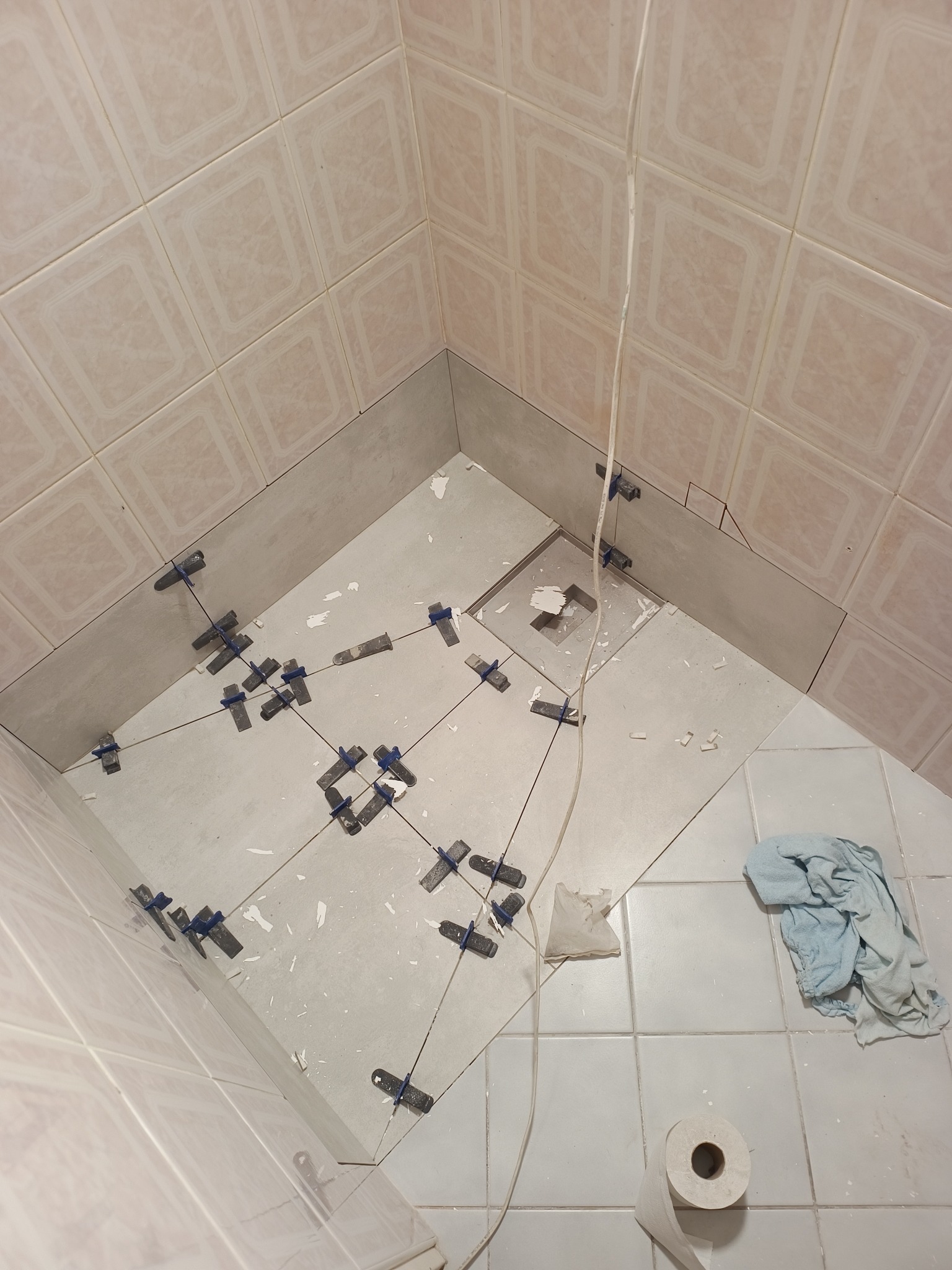 Układanie jasnych płytek w rogu łazienki, widoczne krzyżyki dystansowe, kabel zwisający z góry, ręcznik i rolka papieru toaletowego na podłodze.