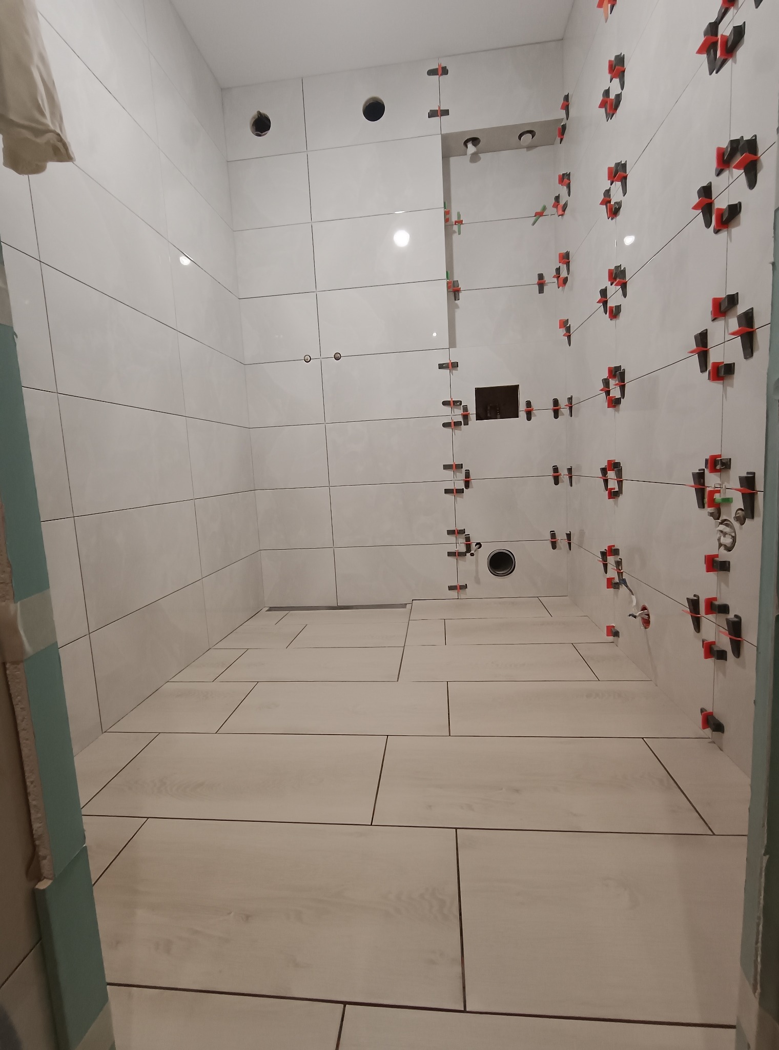 Biała łazienka w trakcie wykańczania. Widoczne płytki na ścianach z systemem poziomowania, ułożone płytki podłogowe imitujące drewno i odpływ liniowy.