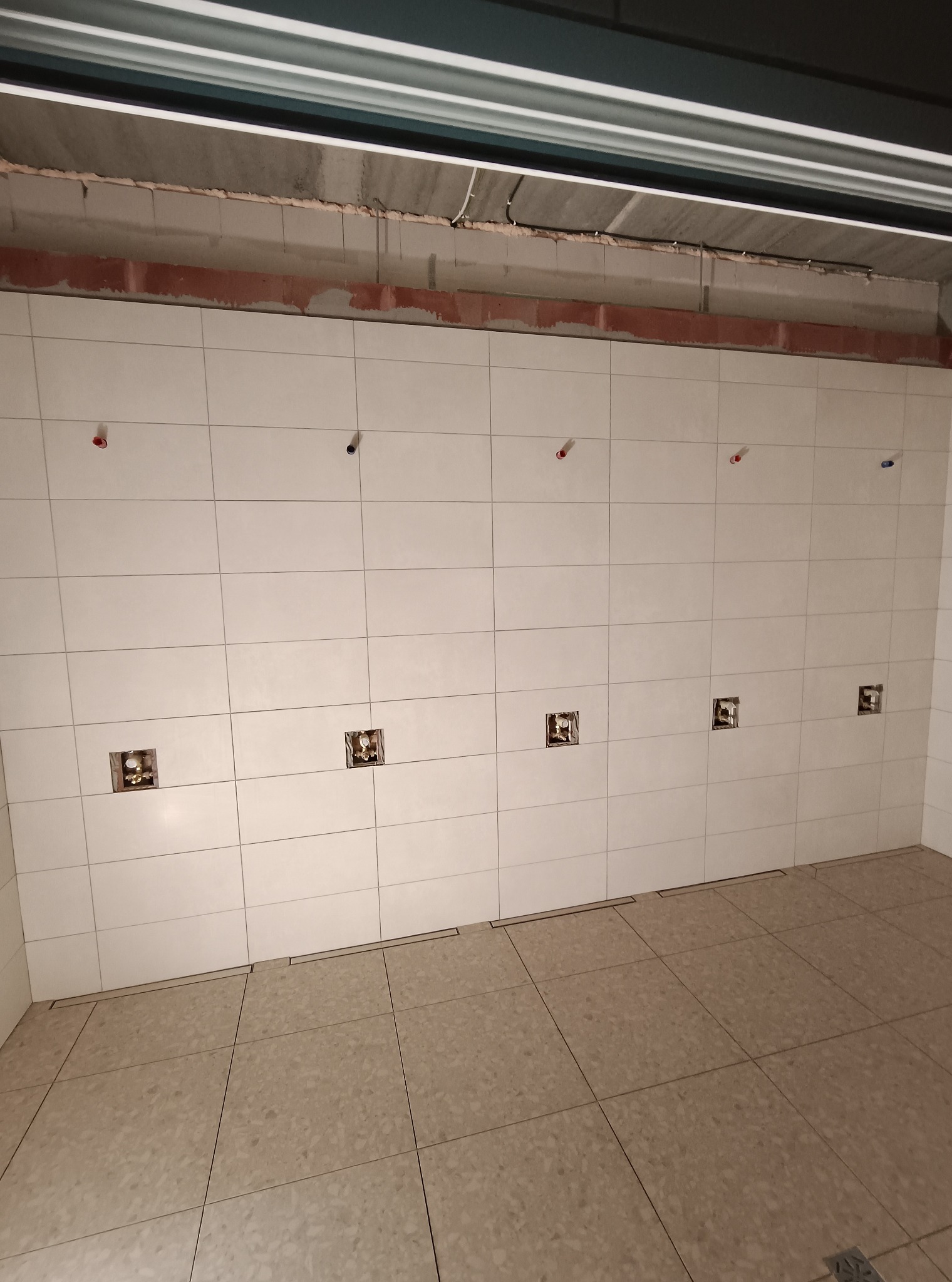 Ściana i podłoga wyłożone jasnymi płytkami. Widoczne otwory na gniazdka i rury. Minimalistyczny design, surowy stan wykończenia.