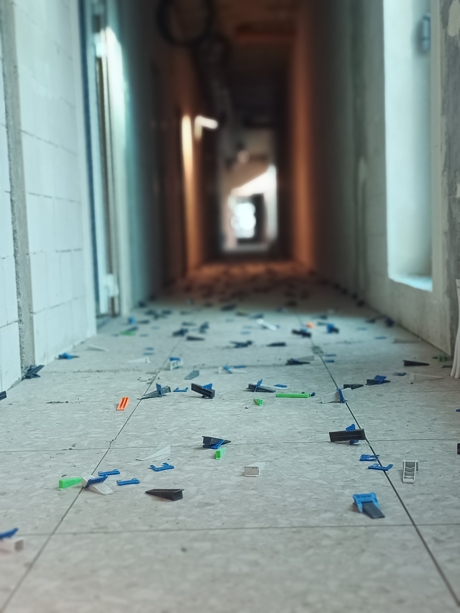 Układanie płytek podłogowych w korytarzu z użyciem klipsów poziomujących. Widok z perspektywy podłogi, płytki w odcieniach szarości, w tle białe ściany i drzwi.