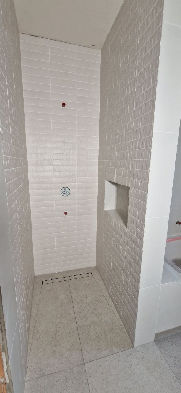 Nowoczesna kabina prysznicowa z jasnymi, prostokątnymi płytkami ułożonymi w cegiełkę, z wnęką na kosmetyki i odpływem liniowym w podłodze.