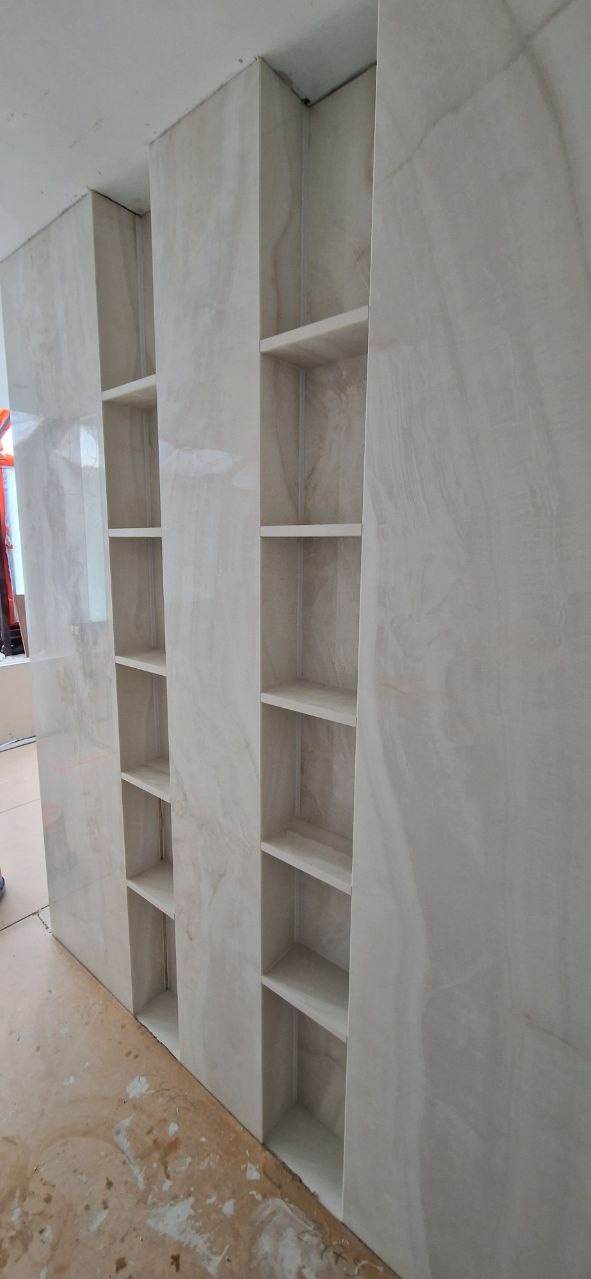 Wykończona wnęka ścienna z półkami, obłożona płytkami imitującymi marmur. Widoczne niedociągnięcia na podłodze sugerują trwające prace wykończeniowe.