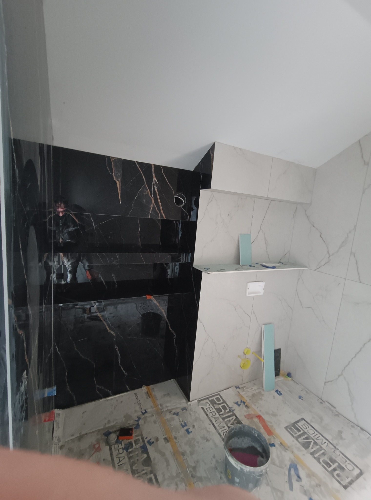 Łazienka w trakcie wykańczania: czarne płytki imitujące marmur na ścianie z wnękami, białe płytki z szarym wzorem, zabezpieczona podłoga, widoczne narzędzia i wiadro.