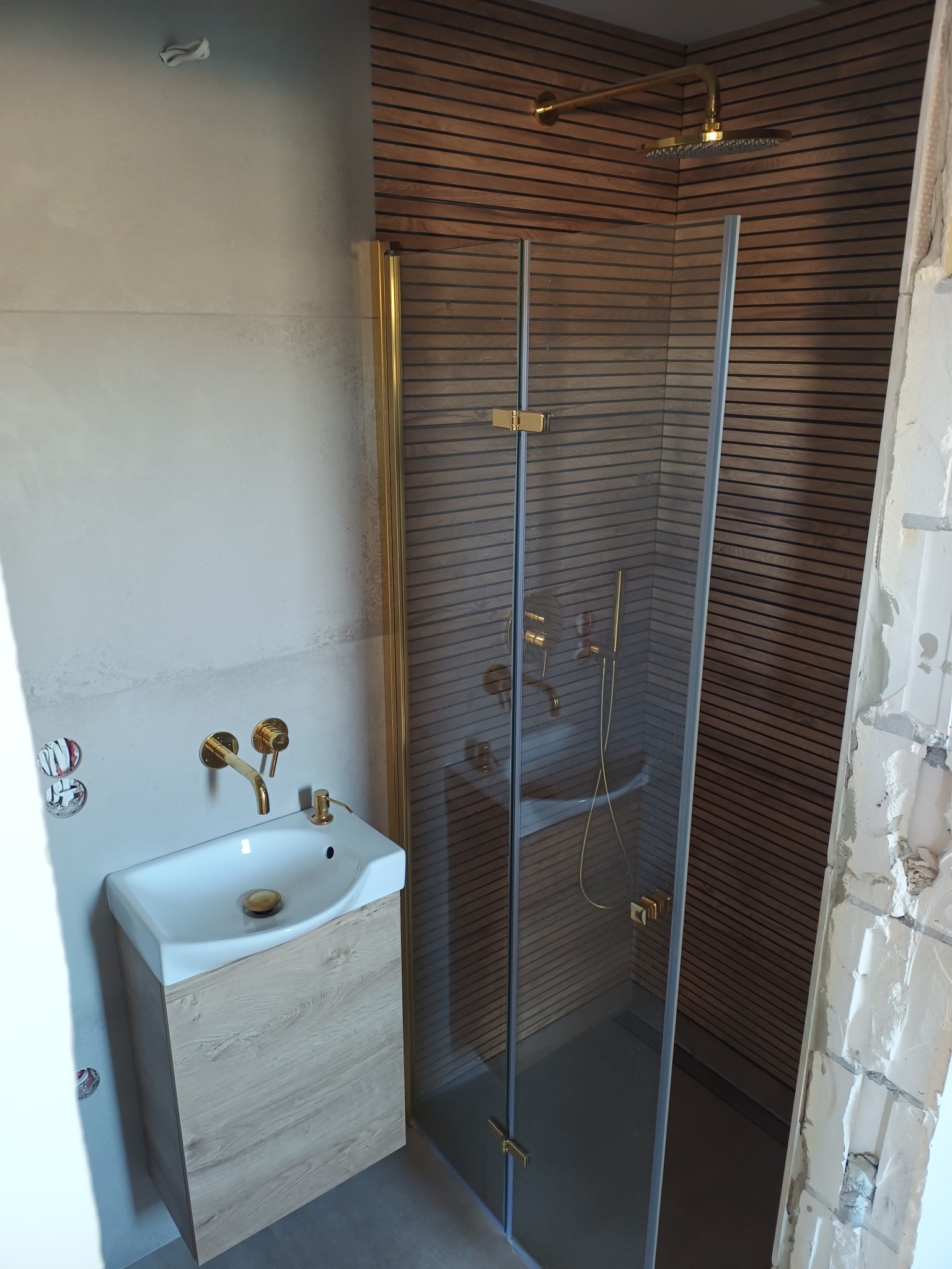 Nowoczesna łazienka z drewnianą okładziną, złotą armaturą i kabiną prysznicową. Umywalka z szafką w jasnym drewnie. Fragment ściany w trakcie wykończenia.