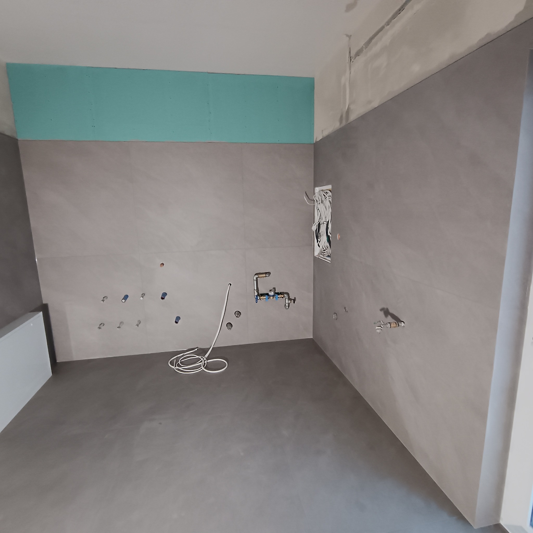 Szara łazienka w trakcie wykańczania: położone płytki, widoczne instalacje wodne i elektryczne, przygotowana ściana pod zabudowę. Minimalistyczny design w surowym stanie.