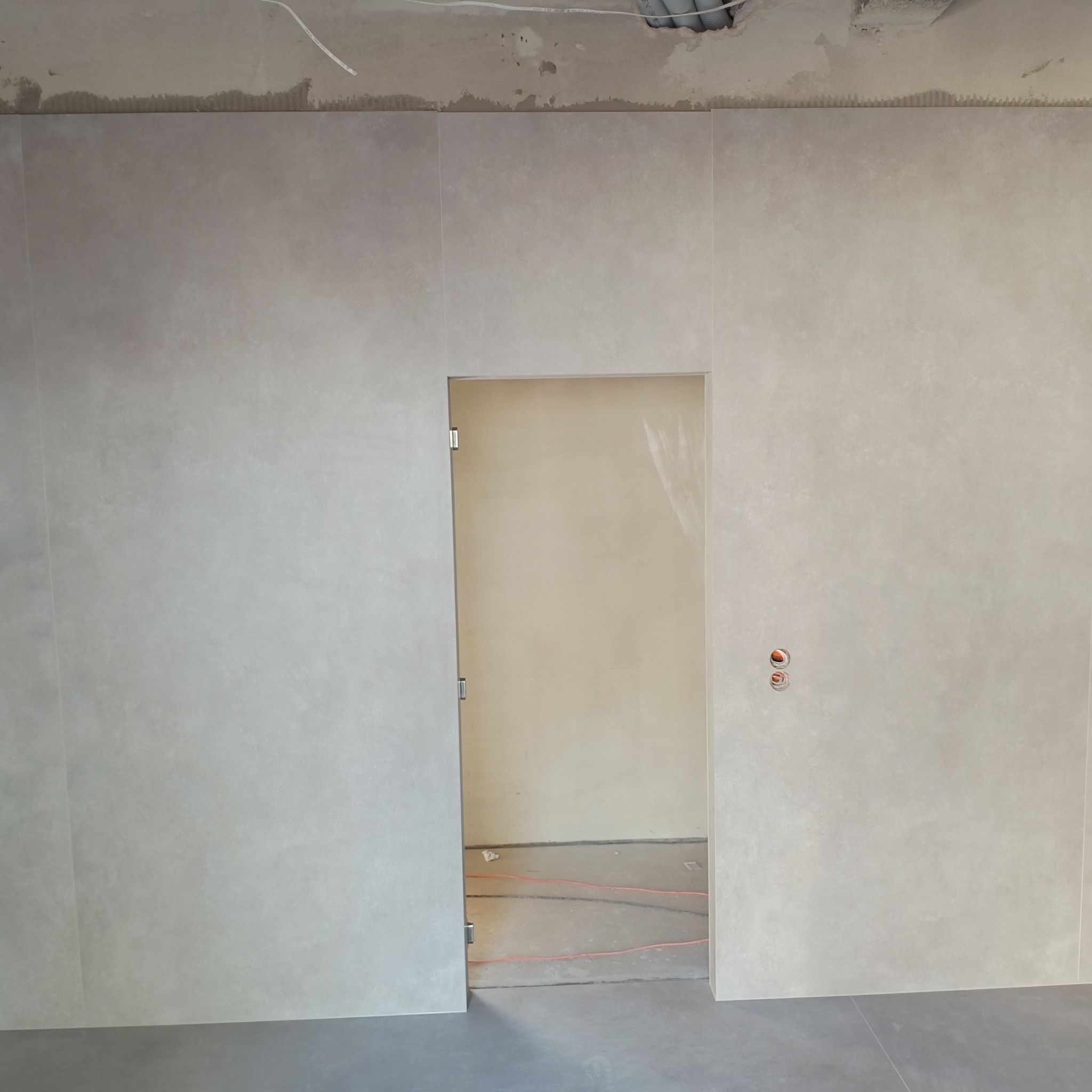 Wykończona ściana w łazience z dużych, beżowych płytek, widoczne gniazdka i fragment instalacji podtynkowej. W tle widoczne ogrzewanie podłogowe.