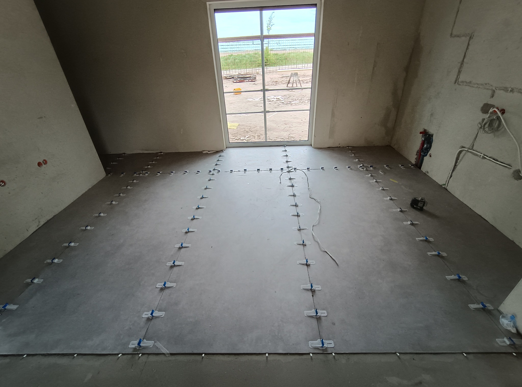 Szare płytki podłogowe ułożone w rzędach z użyciem systemu poziomowania, widoczne białe klipsy i kliny. W tle okno i surowe ściany w trakcie prac wykończeniowych.