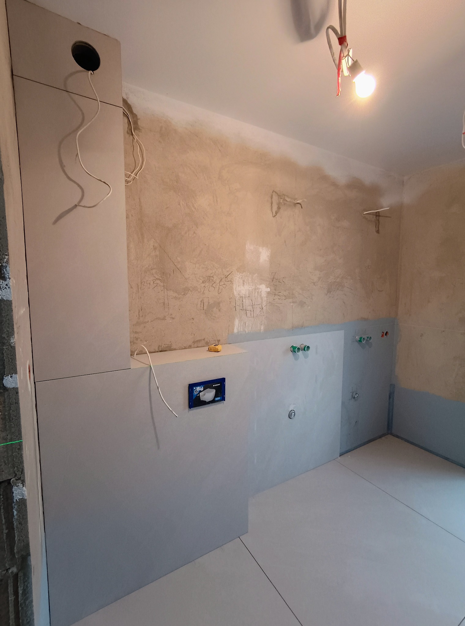 Wnętrze łazienki w trakcie prac wykończeniowych: ułożone płytki podłogowe i częściowo ścienne, widoczne instalacje wodne i elektryczne, surowa ściana w tle.