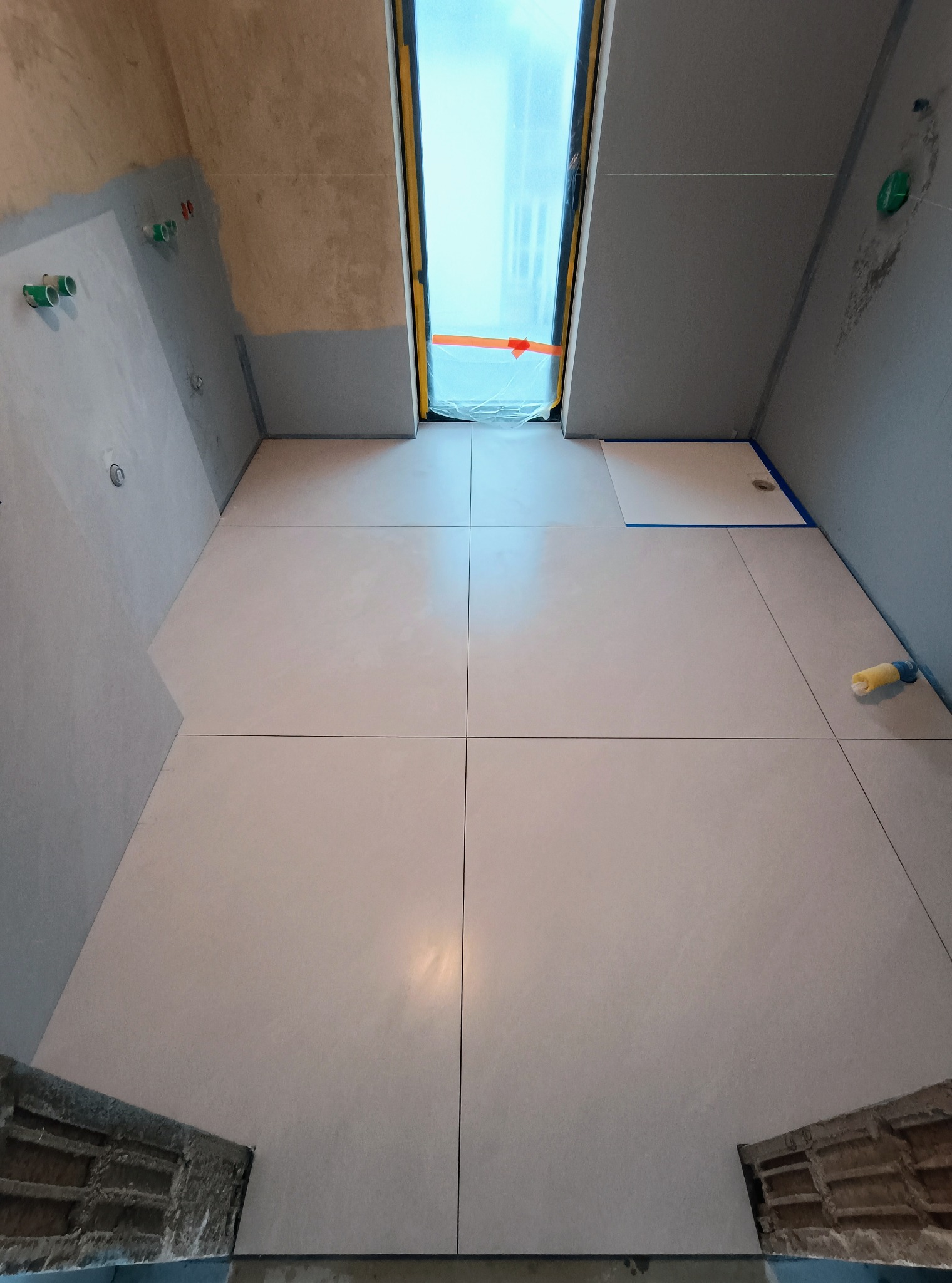 Jasne, kwadratowe płytki podłogowe w trakcie układania w łazience z oknem dachowym. Widoczne przygotowane ściany i instalacje.