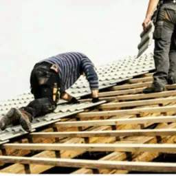 Dwóch dekarzy montuje szare dachówki na drewnianej konstrukcji dachu. Jeden z nich klęczy, drugi stoi trzymając dachówkę. Widok z dołu na jasne niebo.