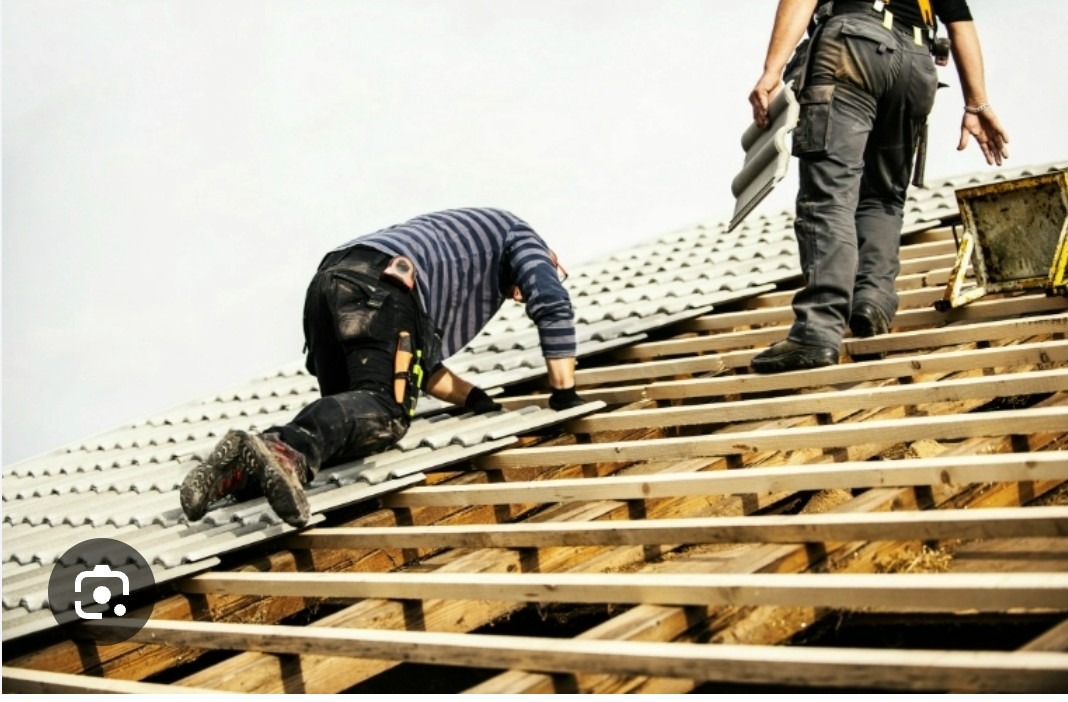 Dwóch dekarzy montuje szare dachówki na drewnianej konstrukcji dachu. Jeden z nich klęczy, drugi stoi trzymając dachówkę. Widok z dołu na jasne niebo.