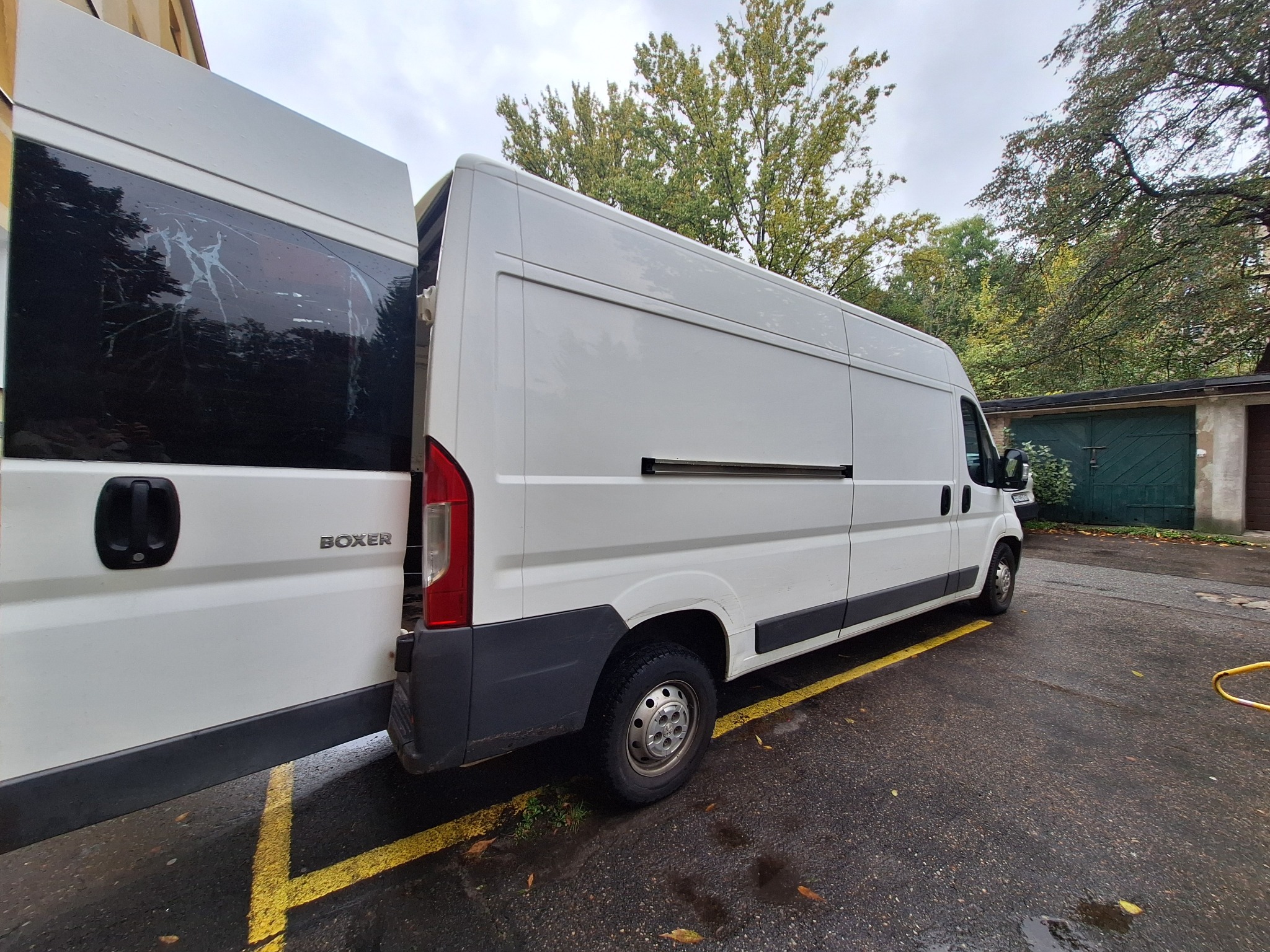 Biały furgon marki Boxer z otwartymi drzwiami, zaparkowany na mokrym asfalcie z żółtymi liniami, widok z boku na tle drzew i garażu.