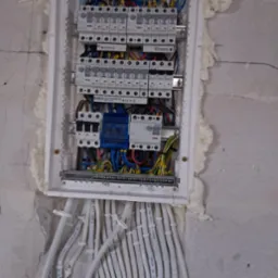 Rozdzielnica elektryczna w trakcie montażu, widoczne kable z oznaczeniami, zabezpieczenia i pianka montażowa wokół obudowy. Przewody ułożone pionowo.