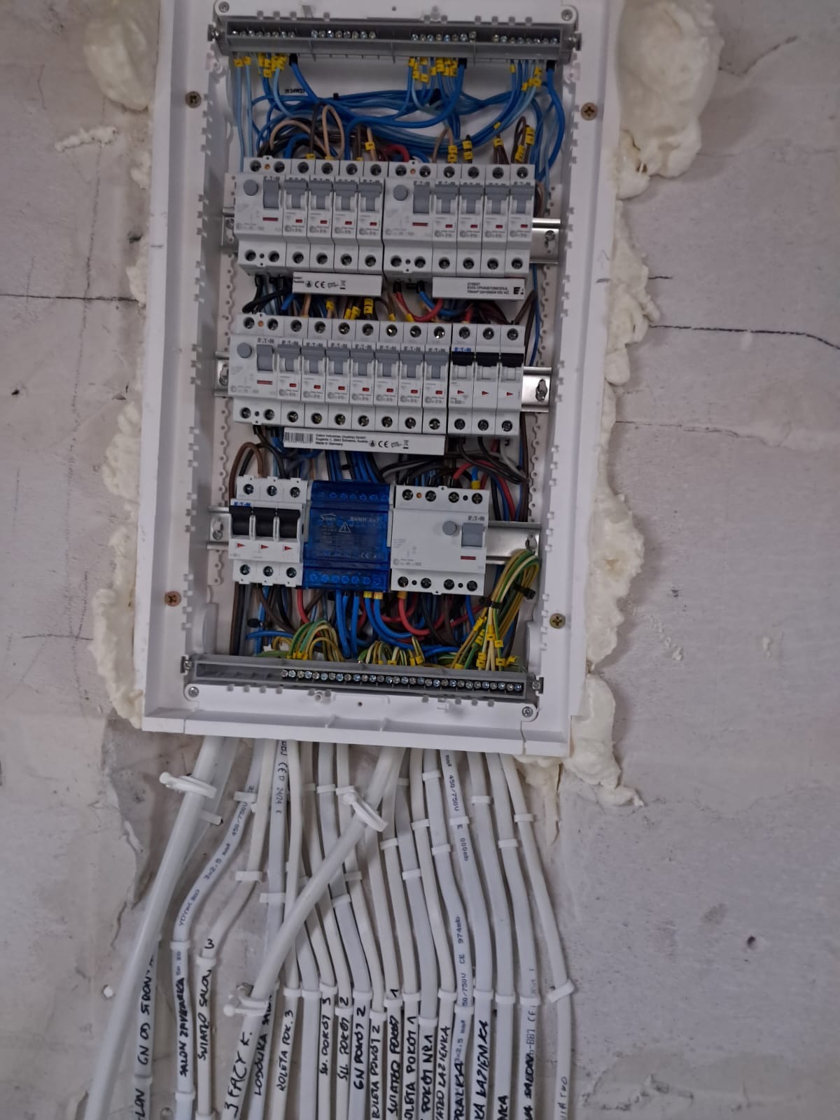 Rozdzielnica elektryczna w trakcie montażu, widoczne kable z oznaczeniami, zabezpieczenia i pianka montażowa wokół obudowy. Przewody ułożone pionowo.