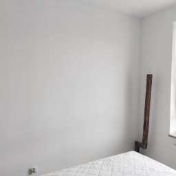 Przemysław Fedak PFD Trans - Jasne, pomalowane na biało wnętrze sypialni z fragmentem łóżka i drewnianym elementem dekoracyjnym przy ścianie obok okna. Widoczny grzejnik pod oknem i gniazdko elektryczne.