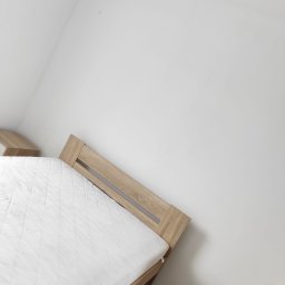 Przemysław Fedak PFD Trans - Świeżo pomalowany pokój z łóżkiem z jasnego drewna i białą pościelą. Minimalistyczne wnętrze po remoncie, białe ściany, brak dekoracji. Kabel na podłodze.