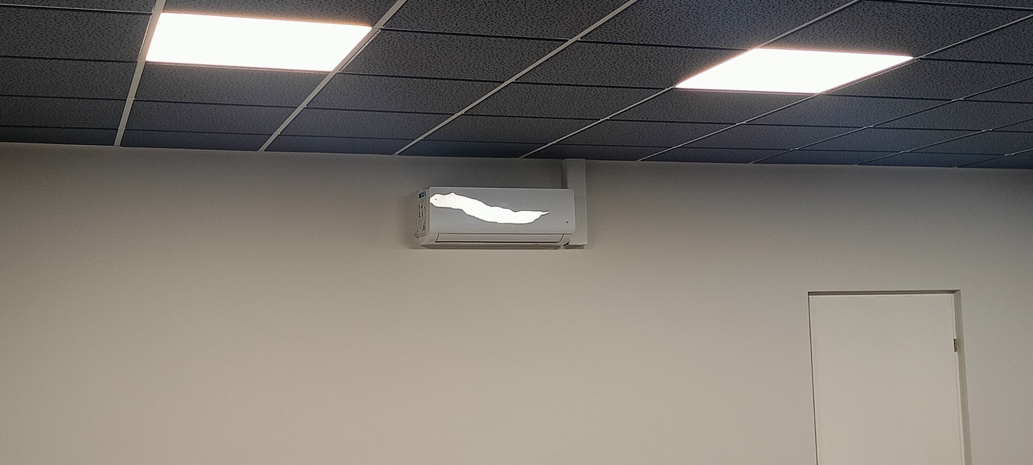 Biała ściana z zamontowanym klimatyzatorem, z widocznym miejscem po usuniętej farbie. Sufit kasetonowy z oświetleniem. Białe drzwi widoczne w tle po prawej stronie.