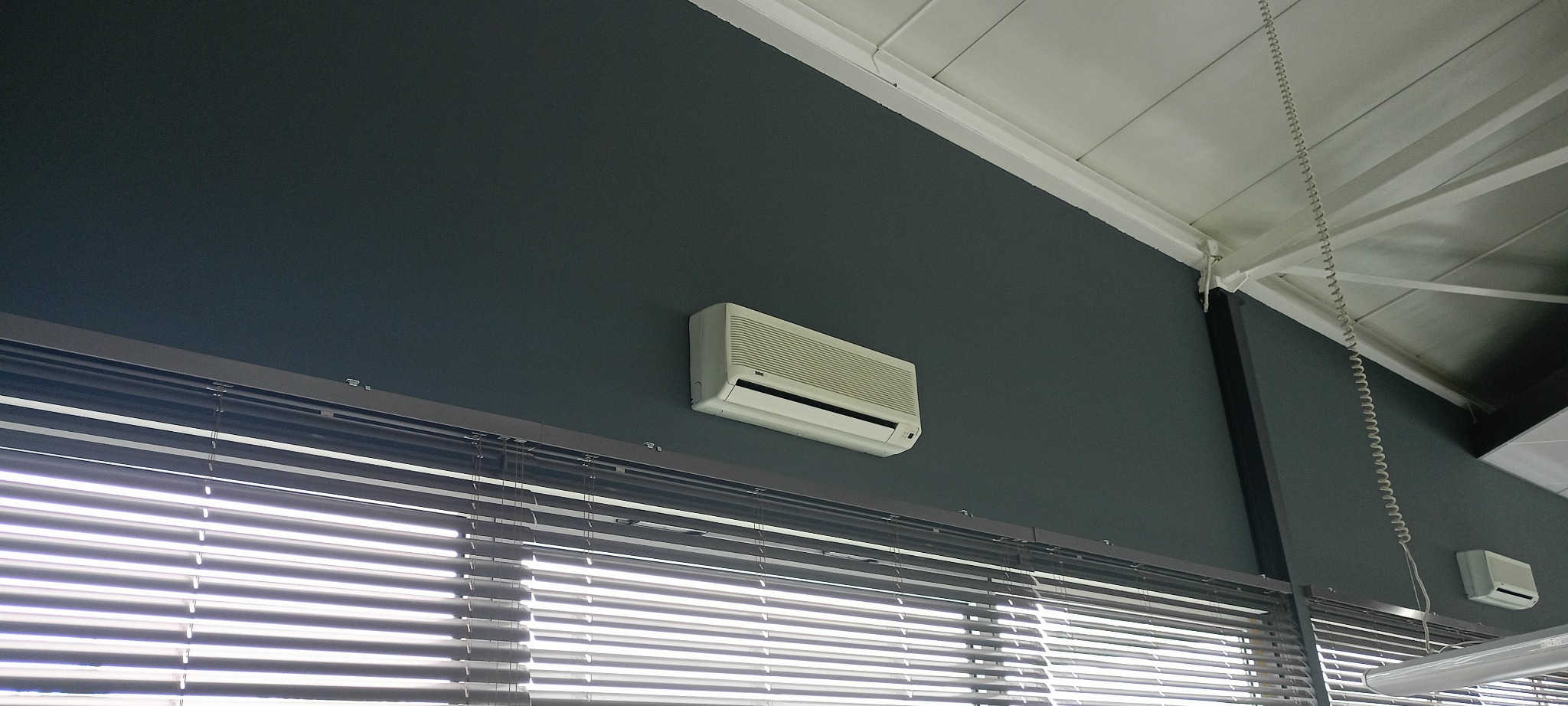 Ściana pomalowana na szaro z zamontowanym klimatyzatorem i żaluzjami. Widoczny fragment sufitu. Świeżo pomalowane ściany w pomieszczeniu biurowym.