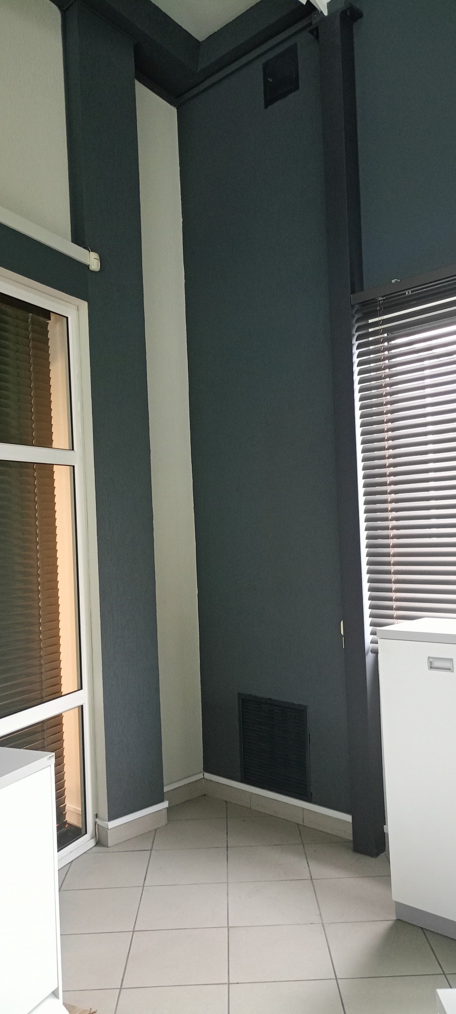 Pomieszczenie po malowaniu: szara ściana, biała kolumna, okno z żaluzjami. Widoczna kratka wentylacyjna i fragment białej szafki. Jasne płytki na podłodze.