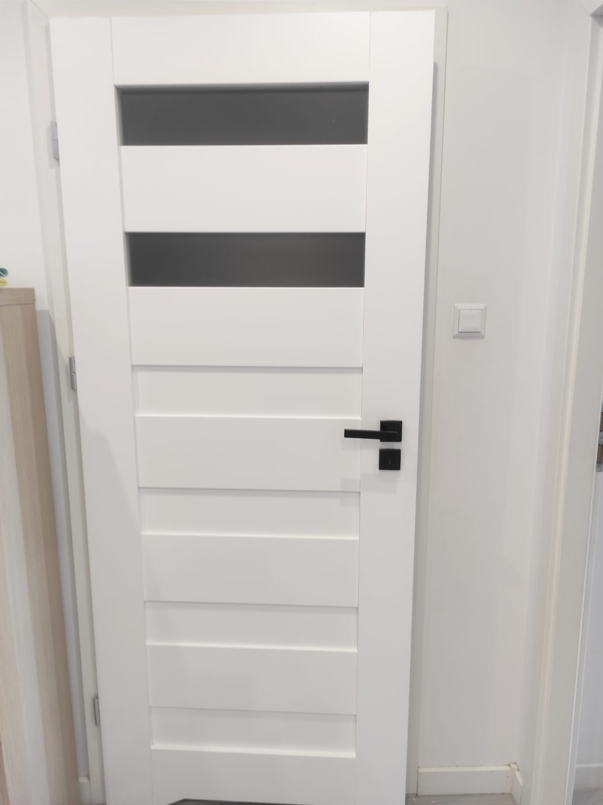 Białe, nowoczesne drzwi wewnętrzne z czarną klamką i przeszkleniami, zamontowane w jasnym pomieszczeniu. Minimalistyczny design.
