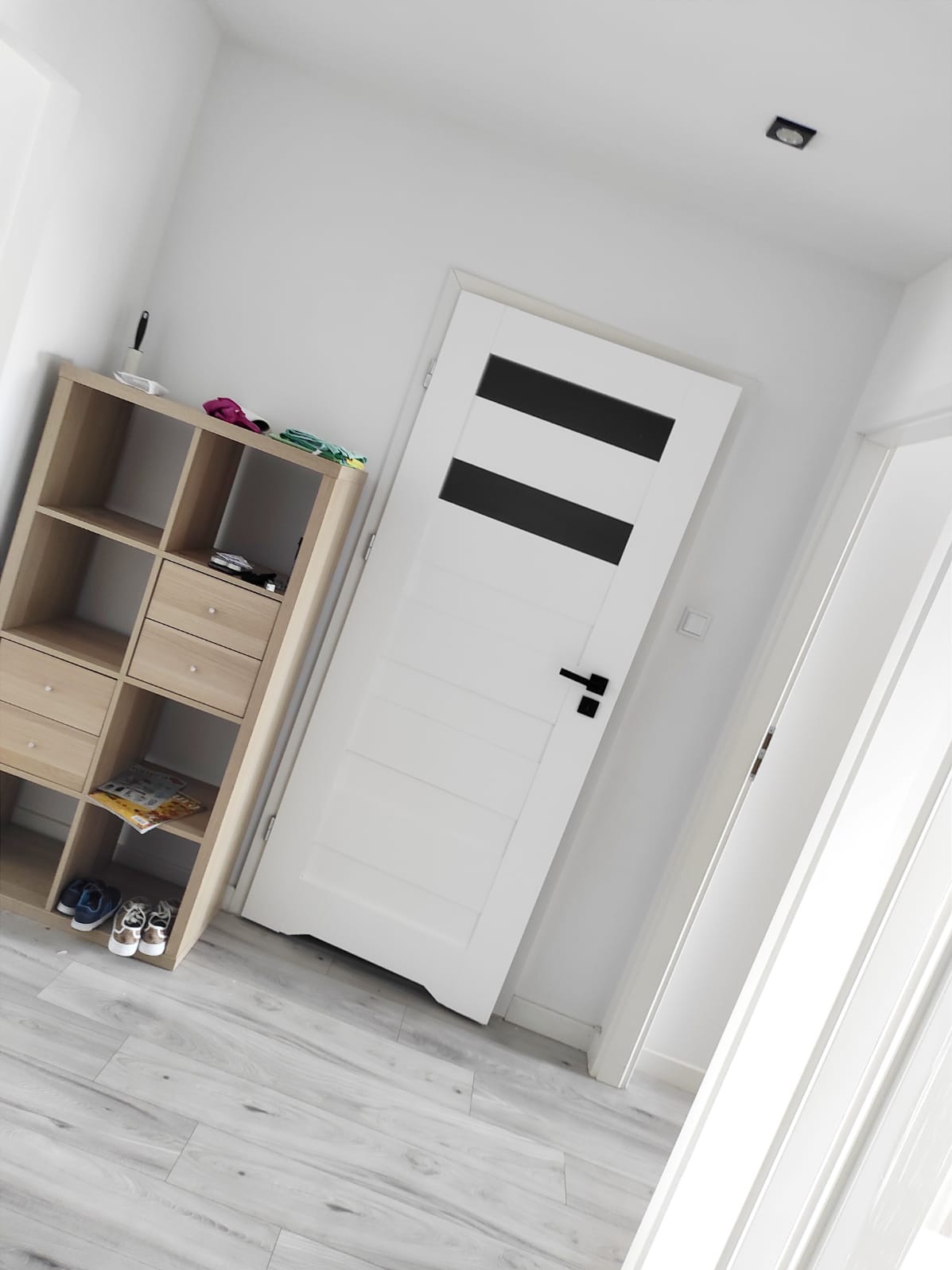 Białe wnętrze: ściana z gładzią, drzwi z czarnymi pasami i regał z butami. Minimalistyczny design, jasne drewno, świeżo po malowaniu.