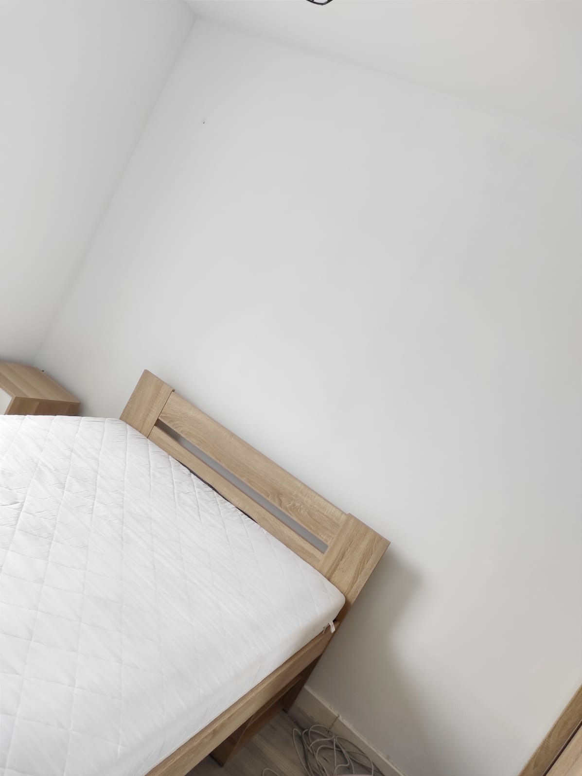 Świeżo pomalowany pokój z łóżkiem z jasnego drewna i białą pościelą. Minimalistyczne wnętrze po remoncie, białe ściany, brak dekoracji. Kabel na podłodze.