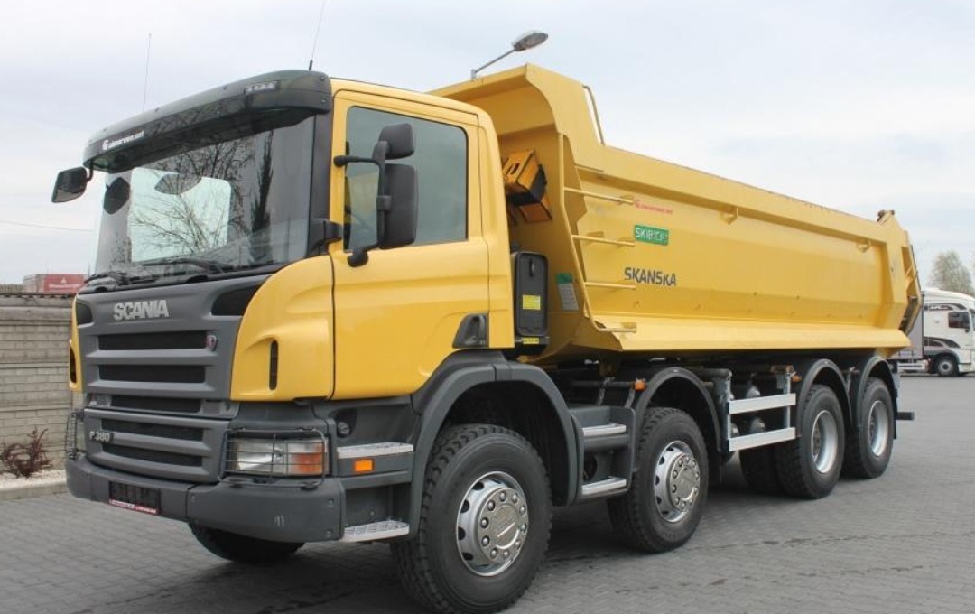 Żółta, sześcioosiowa wywrotka Scania P380 z podniesioną skrzynią ładunkową, zaparkowana na betonowym placu, widok z przodu, lekko z lewej strony.
