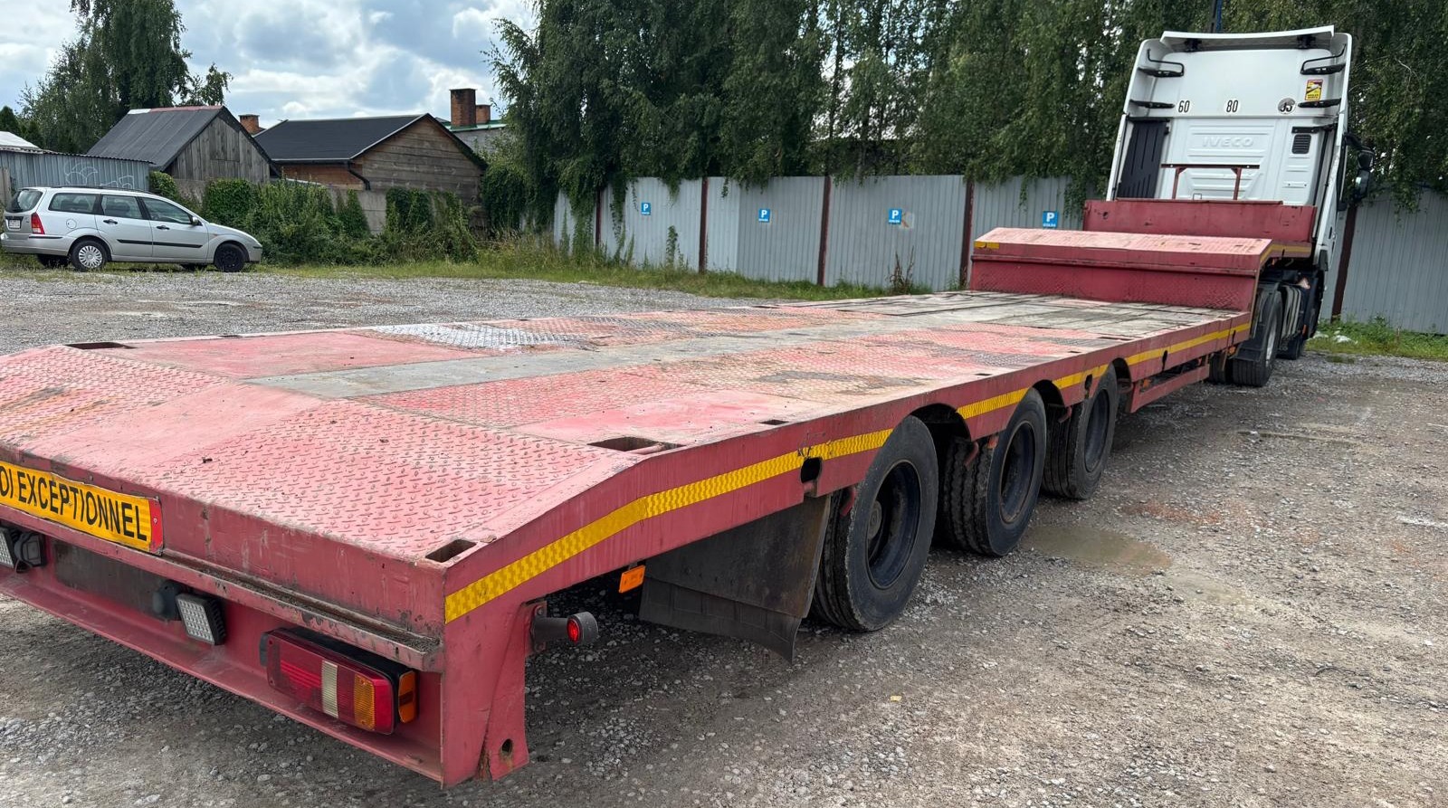 Czerwona, wieloosiowa naczepa niskopodwoziowa na żwirowym podłożu, widok z tyłu, z żółtymi pasami ostrzegawczymi, w tle budynki i zaparkowany samochód.