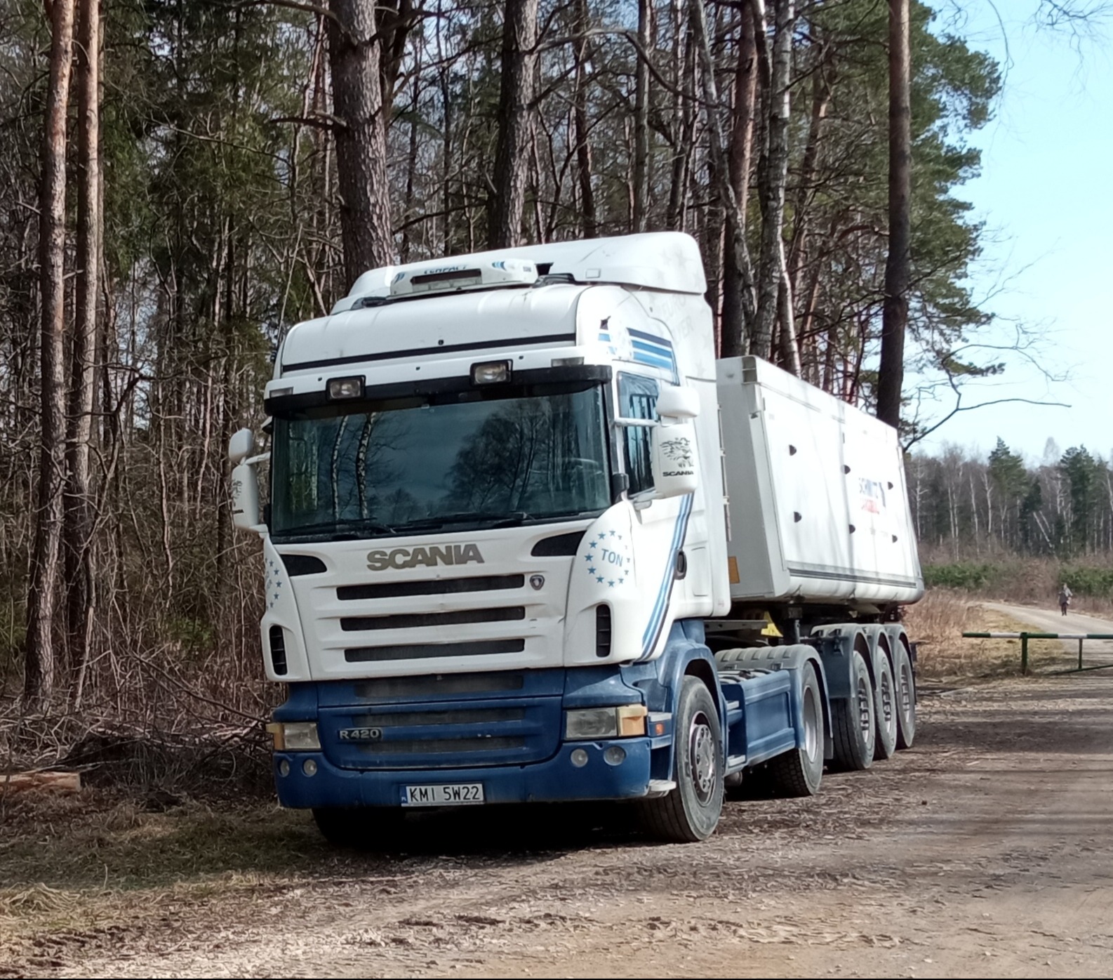 Biało-niebieska ciężarówka Scania R420 z naczepą stoi na leśnej drodze. Widoczne drzewa w tle i osoba w oddali. Rejestracja KMI 5W22.