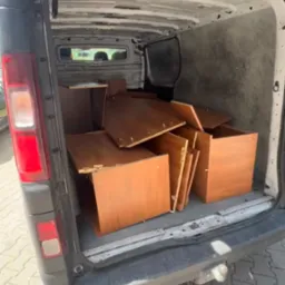 Wnętrze busa wypełnione elementami mebli w odcieniach brązu, przygotowanych do transportu. Widoczne płyty, półki i elementy konstrukcyjne.