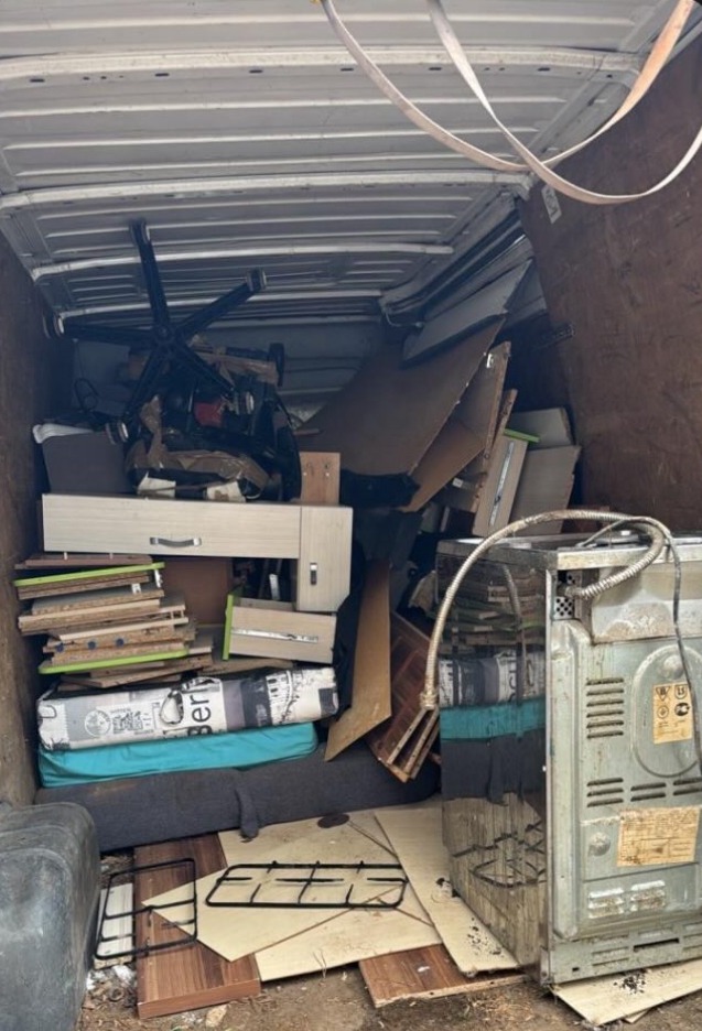 Wnętrze furgonetki wypełnionej meblami, kartonami i sprzętem AGD, sugerujące opróżnianie pomieszczeń. Widoczny fotel biurowy, szafka i kuchenka.