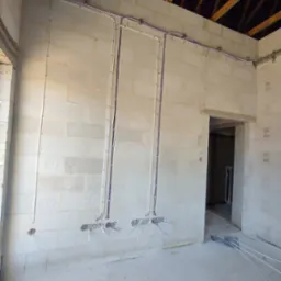 Surowe wnętrze z instalacją elektryczną w białych rurkach na ścianie z bloczków. Widoczne przewody i otwory montażowe. Elementy mocujące na suficie.