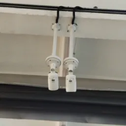 Dwa identyczne, białe czujniki dymu zamontowane równolegle do sufitu, z czarnymi kablami biegnącymi po jego powierzchni. Widoczne mocowania i fragment instalacji.