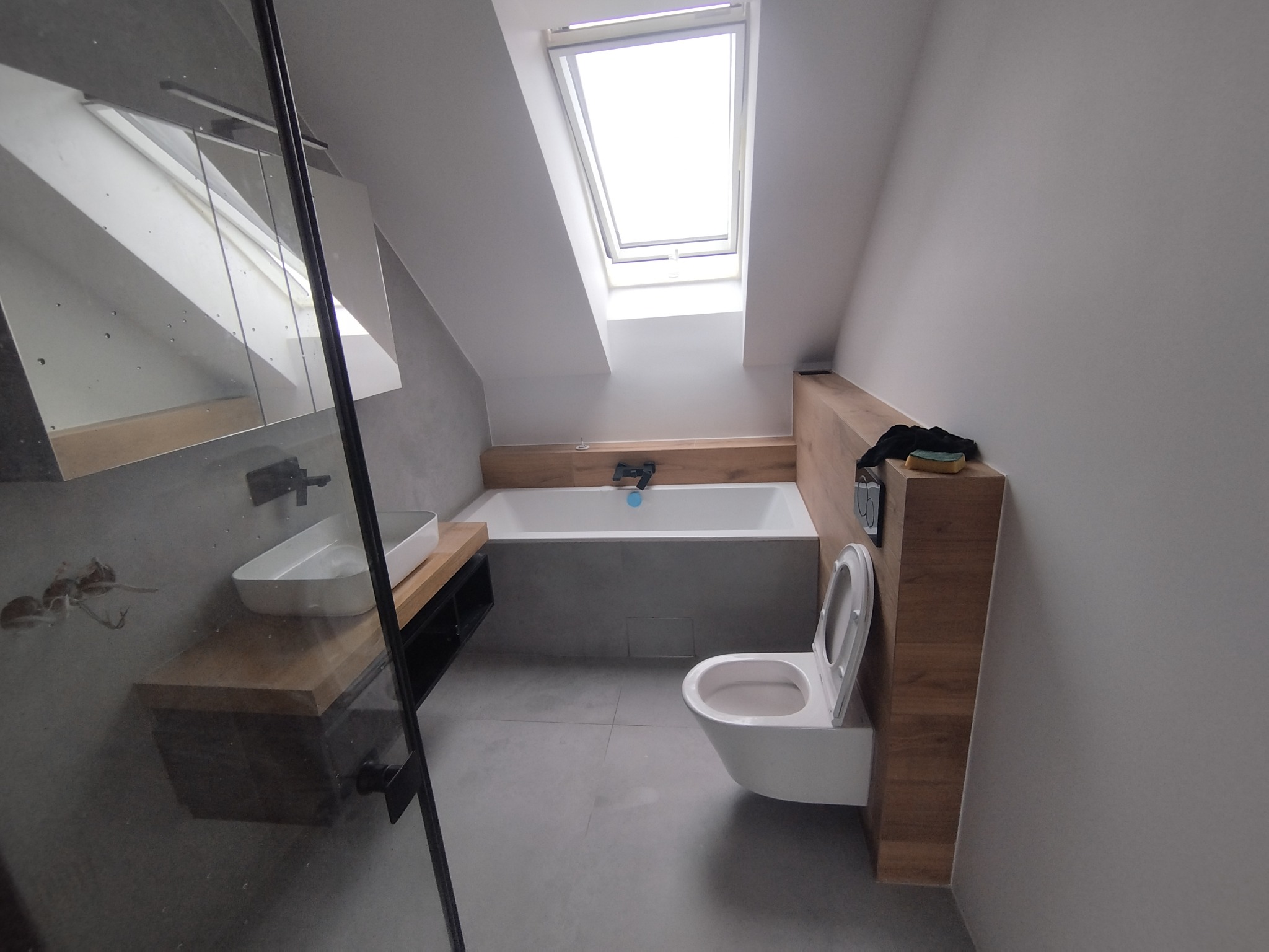 Nowoczesna łazienka na poddaszu w Łodzi z oknem dachowym, szarą glazurą i drewnianymi akcentami. Umywalka nablatowa, toaleta podwieszana.