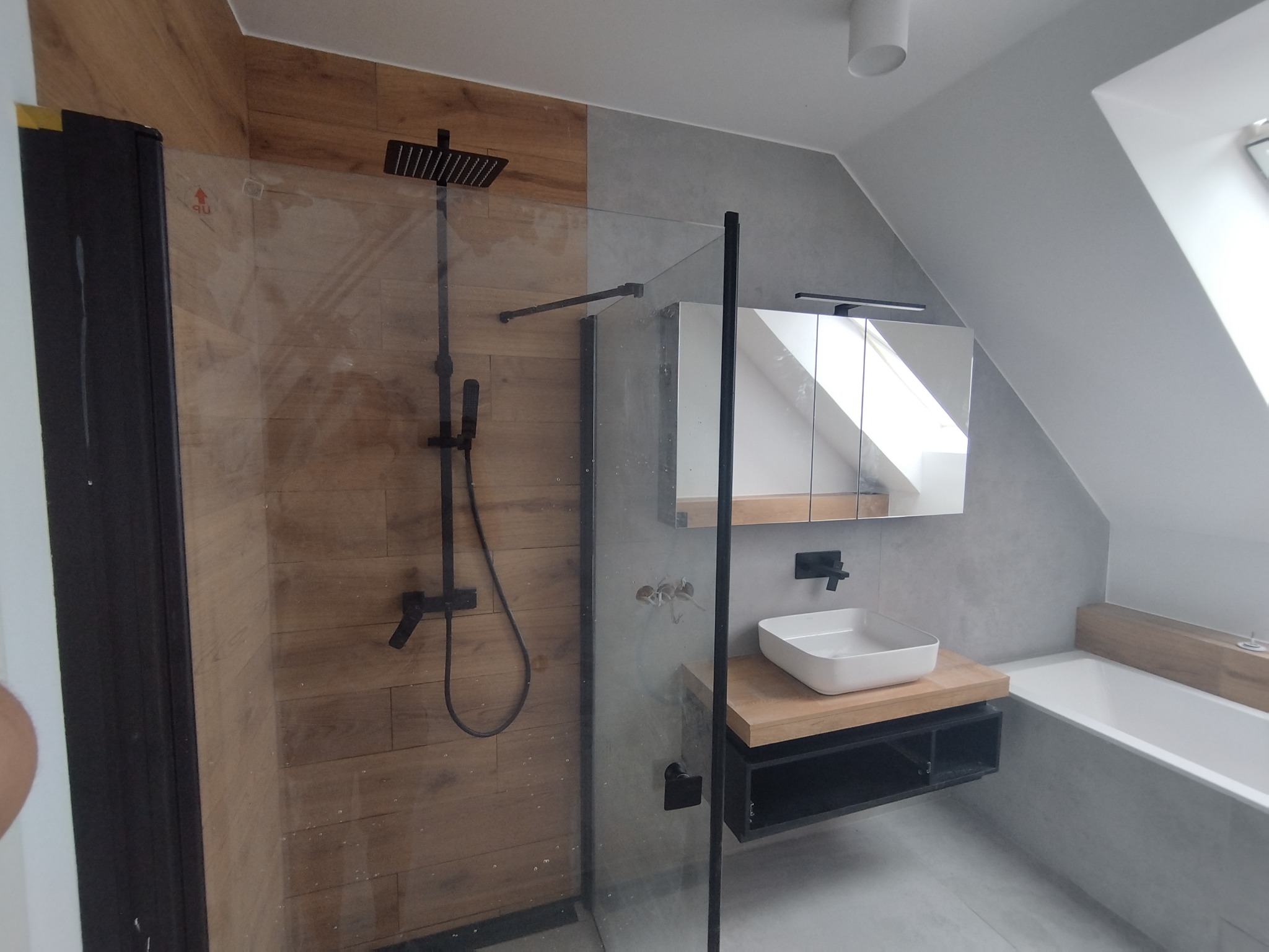 Nowoczesna łazienka na poddaszu: drewnopodobne płytki w kabinie prysznicowej kontrastują z szarą armaturą i białą umywalką na drewnianym blacie.