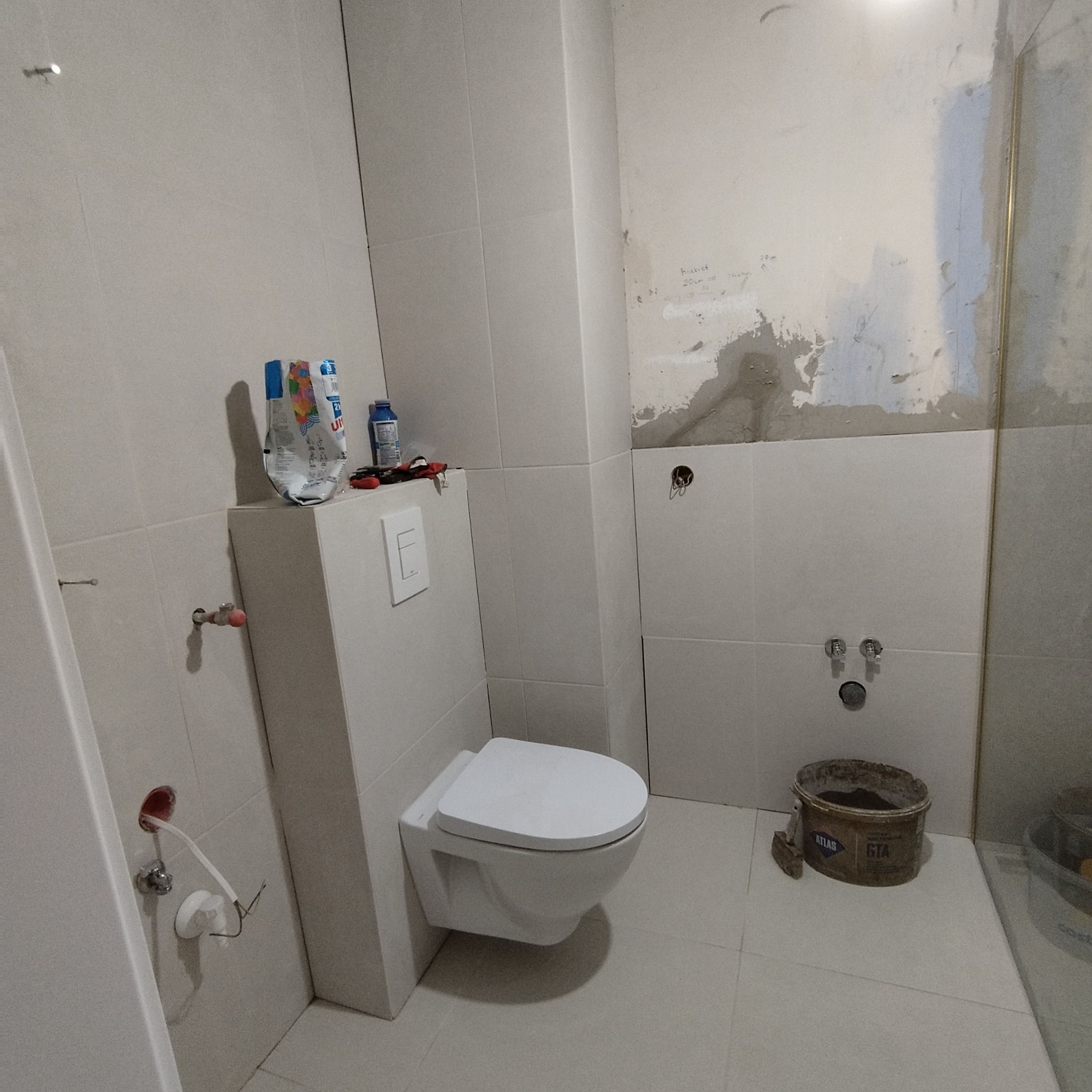 Ściana z częściowo położonymi białymi płytkami w łazience, widoczny podtynkowy stelaż WC, wiadro z zaprawą i narzędzia. Etap prac wykończeniowych.