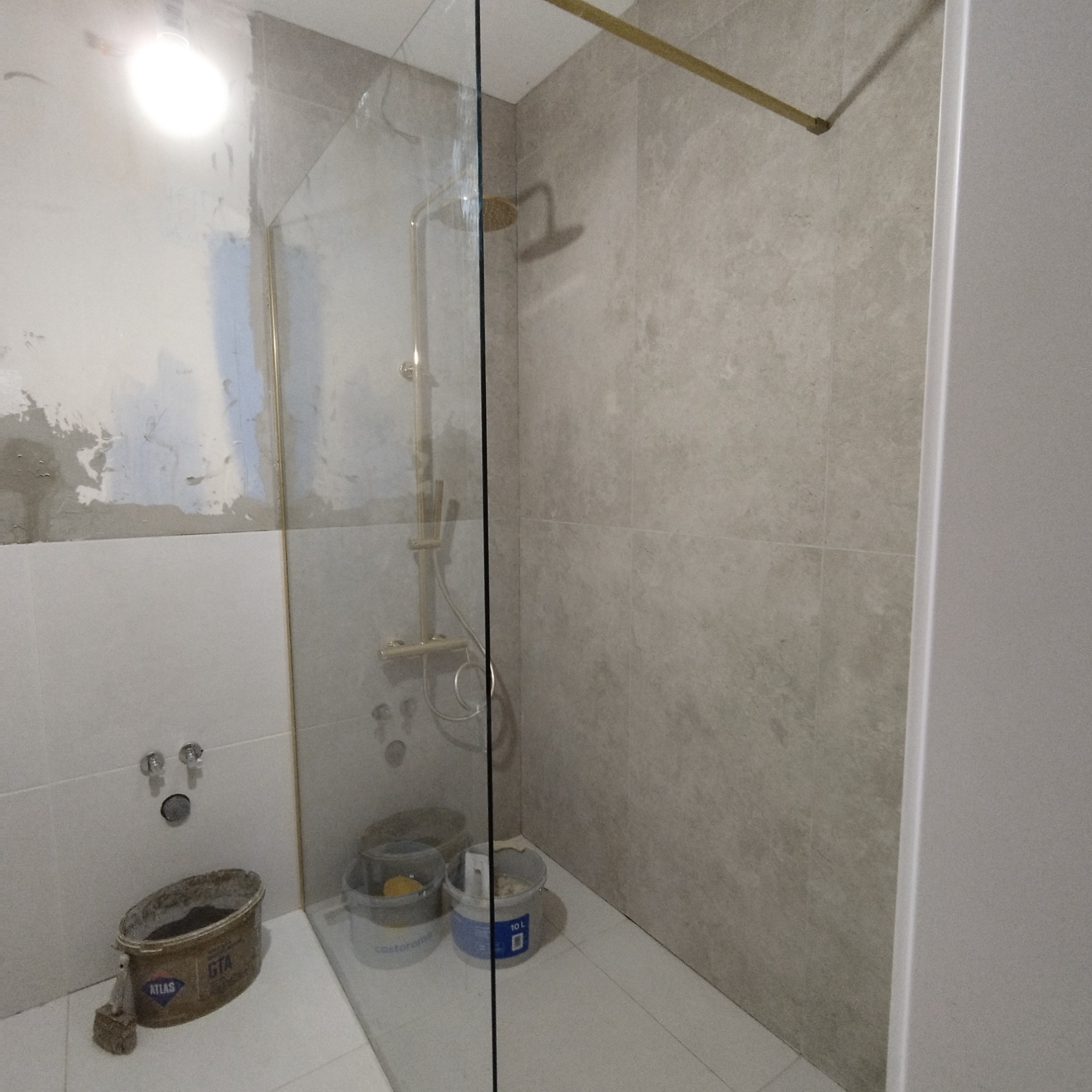 Remont łazienki w toku: złoty zestaw prysznicowy za szklaną ścianką, częściowo wykończone płytki, widoczne wiadra z zaprawą.