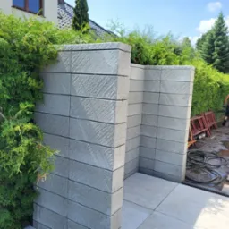 Nowoczesny mur oporowy z szarej kostki, oddzielający strefy ogrodu. W tle zieleń i pracownik w trakcie pracy. Widoczna linia drenażowa i płyty chodnikowe.