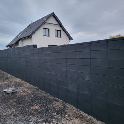 BRUKIO MICHAŁ JABŁOŃSKI - Długi, grafitowy mur z prefabrykatów betonowych, oddzielający posesję z domem o białej elewacji i ciemnym dachu. Na pierwszym planie gruz i elementy muru.