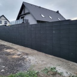 BRUKIO MICHAŁ JABŁOŃSKI - Nowoczesne ogrodzenie z szarej, modułowej kostki betonowej na tle domu z ciemną dachówką i niebem. Minimalistyczny design i proste linie.
