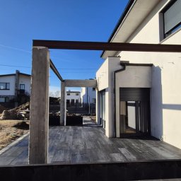 BRUKIO MICHAŁ JABŁOŃSKI - Nowoczesny taras w trakcie budowy: szare deski, betonowe słupy i stalowe belki. W tle dom z białymi ścianami i niezagospodarowany teren.