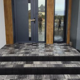 BRUKIO MICHAŁ JABŁOŃSKI - Schody wejściowe z kostki brukowej w odcieniach szarości i czerni, prowadzące do nowoczesnego domu z szarymi drzwiami i jasną elewacją. Widoczne odbicie osoby w szybie.