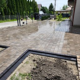 BRUKIO MICHAŁ JABŁOŃSKI - Ułożona kostka brukowa na posesji, z czarną obwódką wokół rabaty. Częściowo mokra nawierzchnia odbija światło. W tle budynek i zieleń.